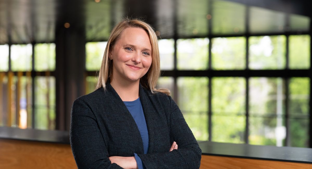Arianna Means stands with arms folded and smiling in a building with glass windows