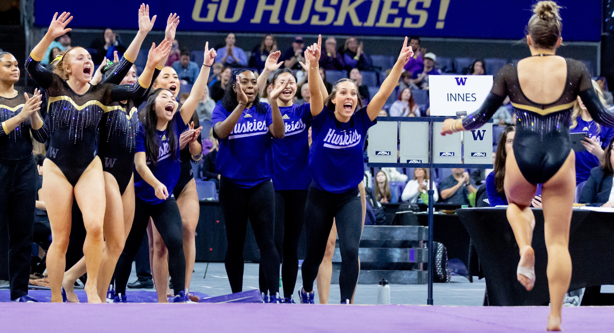 A group of UW gymnasts cheer on the sidelines as a gymnast walks across the mat toward them.