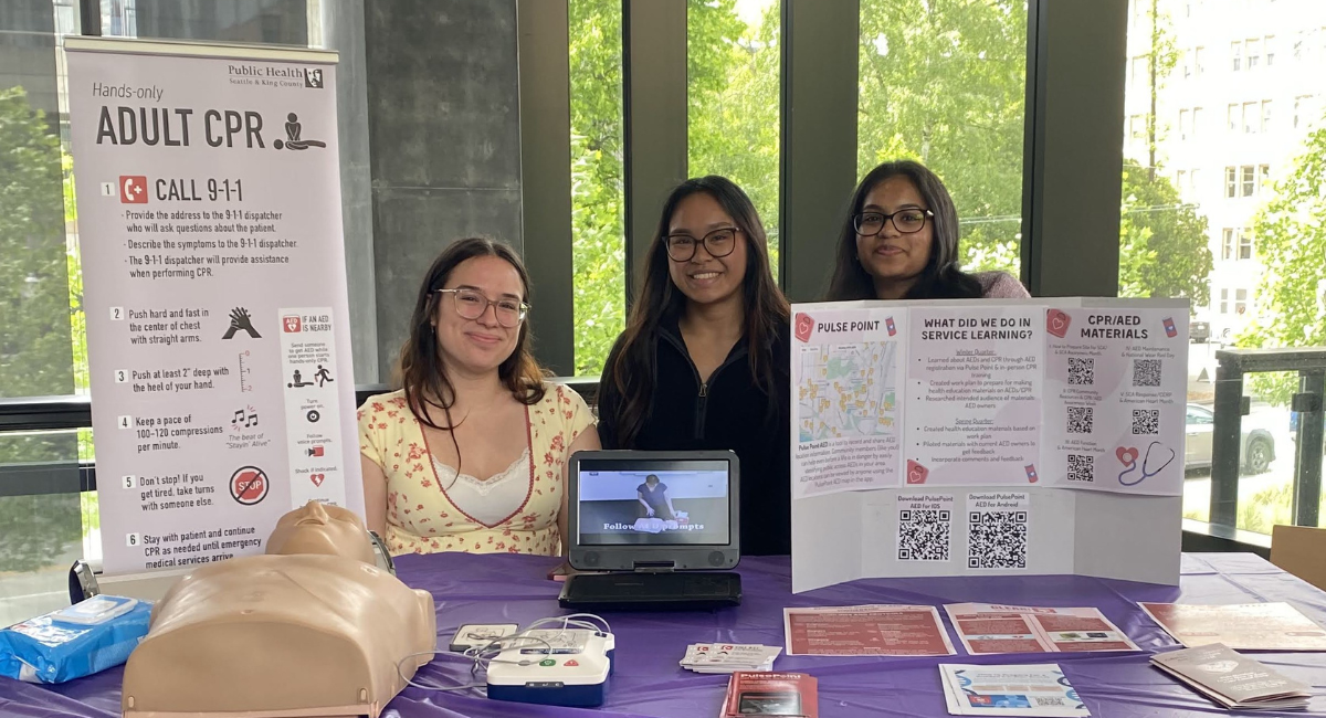 Three students smile as they gather behind table with flyers, posterboard and CPR mannequin 
