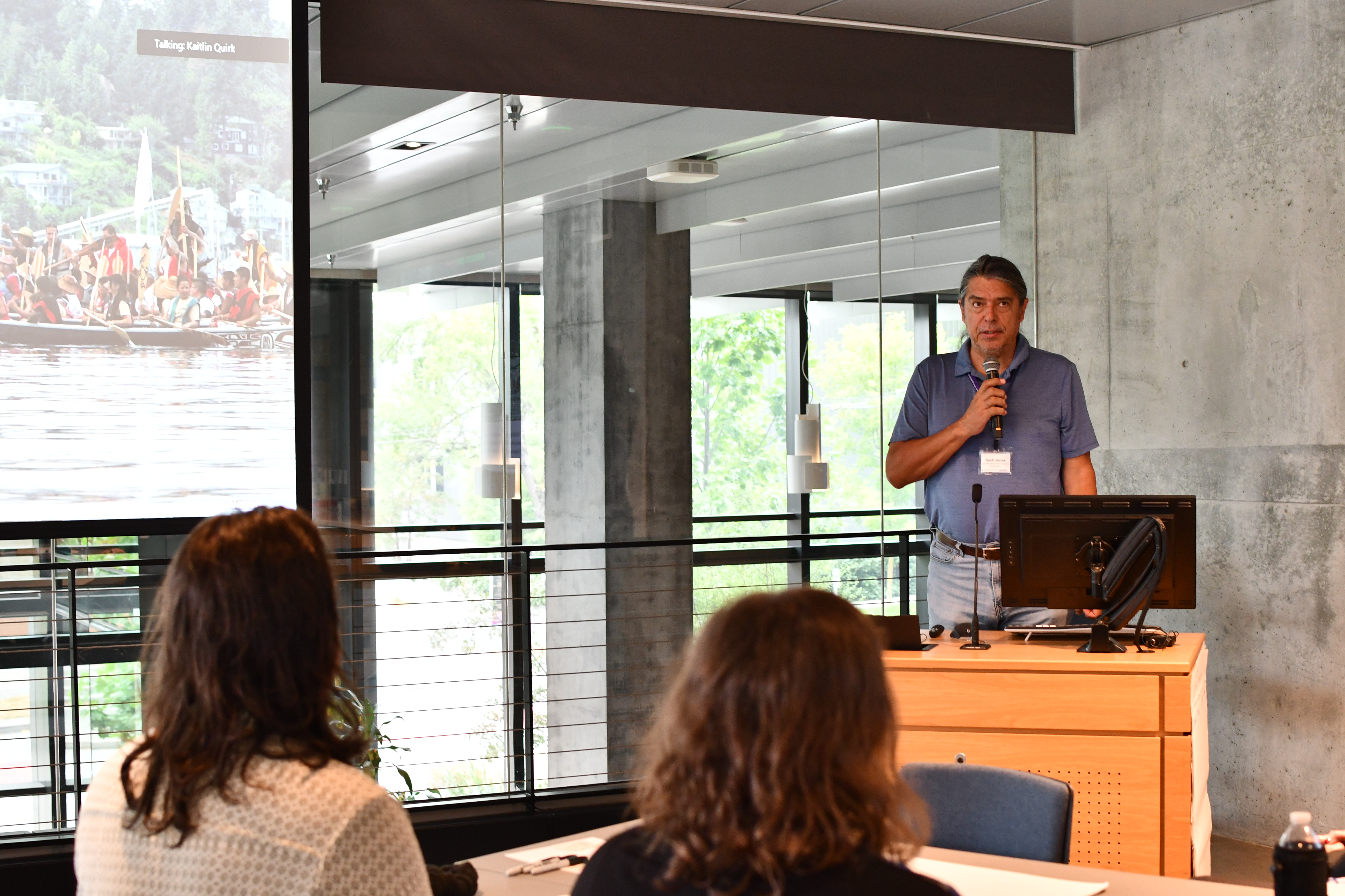 Buck Jones talks into a microphone in a room of people, next to projector of people in canoes.