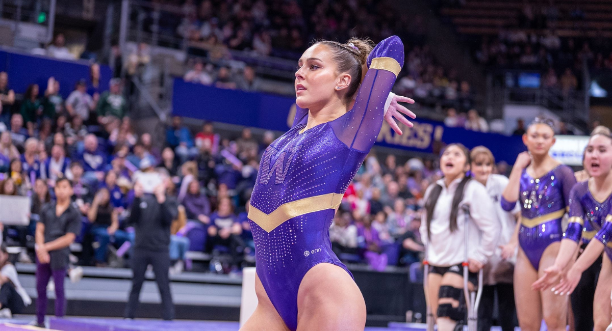 Emily Pires poses during a gymnastics routine with her arms behind her head as teammates cheer behind her.