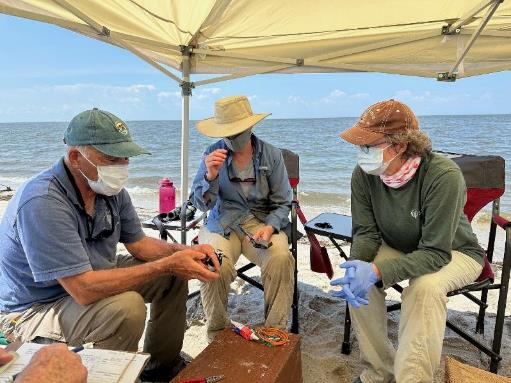 Jeanne wears a mask and surgical gloves while sitting in a folding chair on a beach alongside two other people.