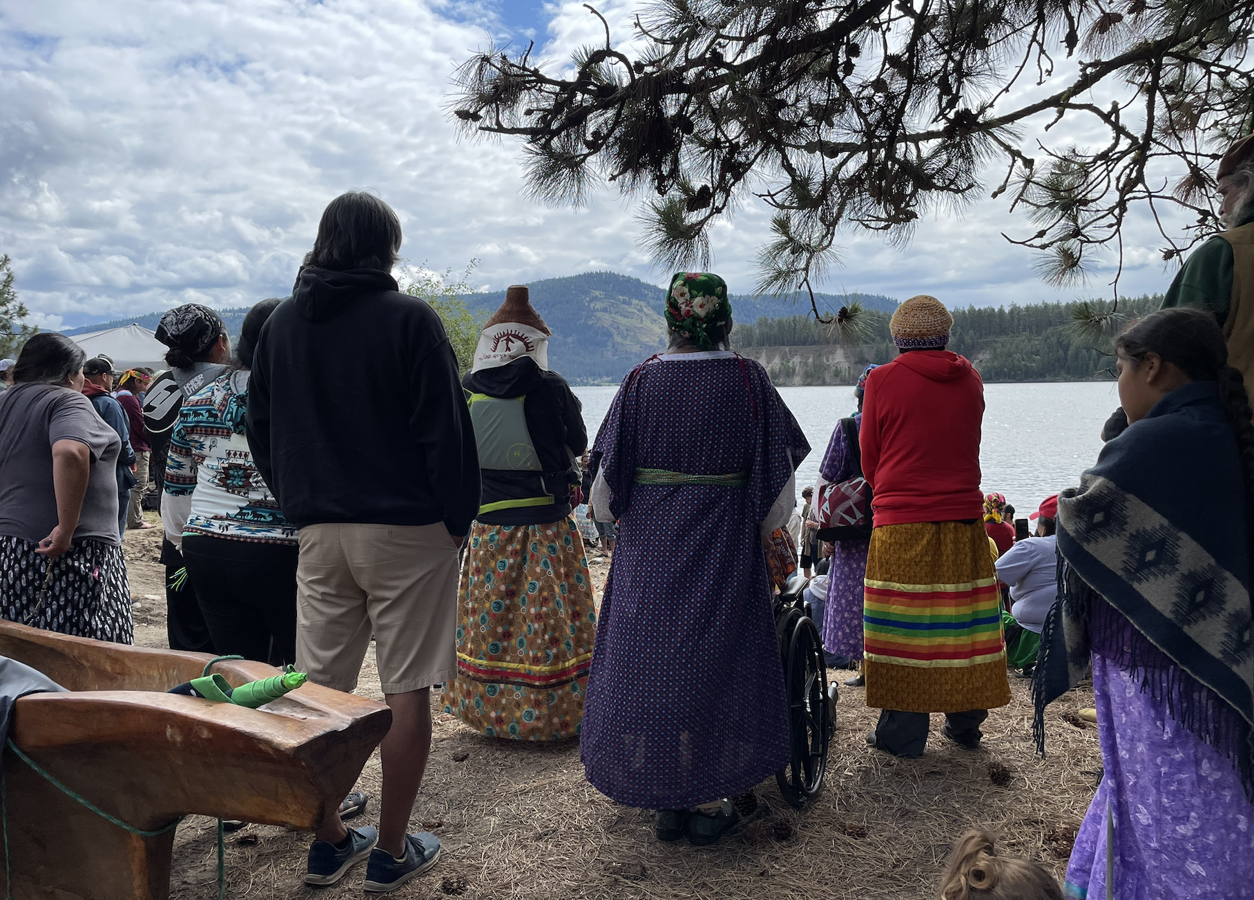 People stand and sit together looking out at a river. A wooden canoe is in the foreground.