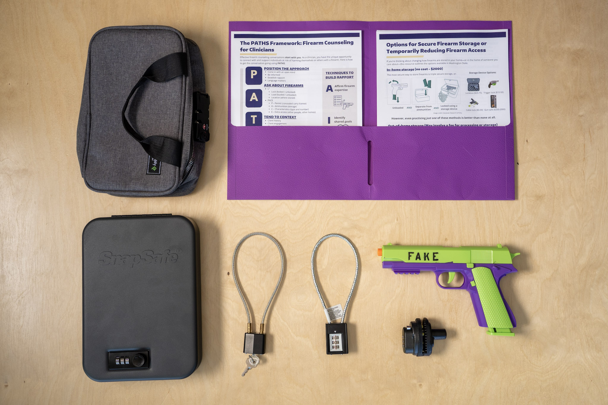 A lockbox, briefcase, two gun locks, a fake firearm and a folder with info pamphlets sit on a table.