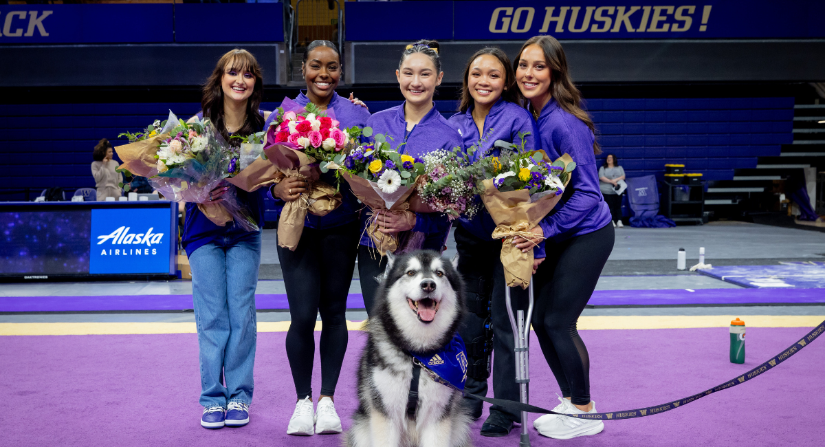 Five members of the gymnastics team stand together in a gym smiling and holding flowers while Dubs the Husky sits in front of them.