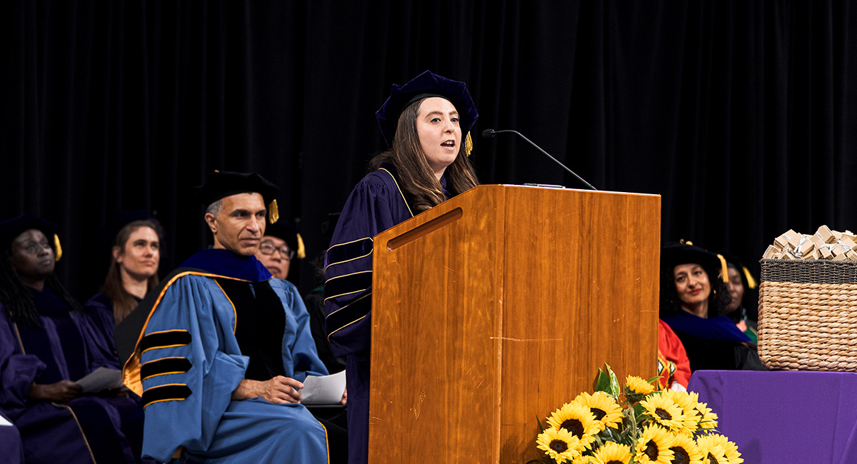 Betty stands at podium on a stage with faculty sitting behind her