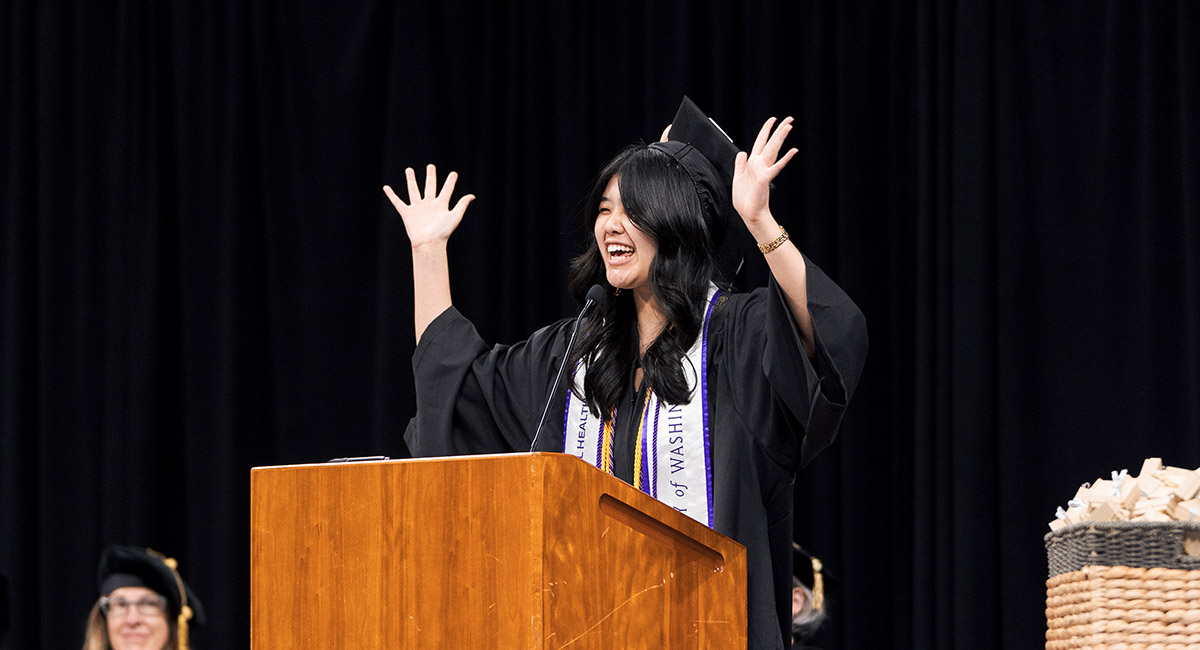 Daphne stands behind a podium with hands in the air, smiling in graduation gown and hat